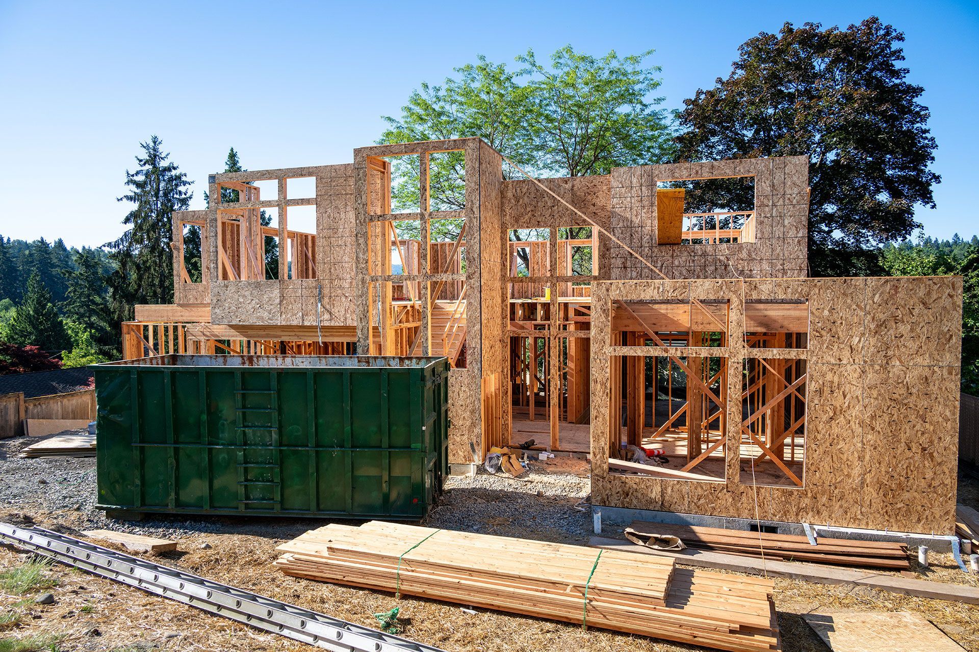 A two-story house under construction with wooden framing. A large green dumpster sits in front of the building.