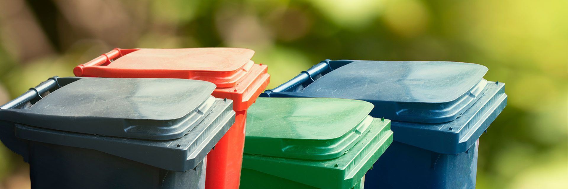 Four colorful trash bins with closed lids of gray, orange, green, and blue, against a blurred green background.