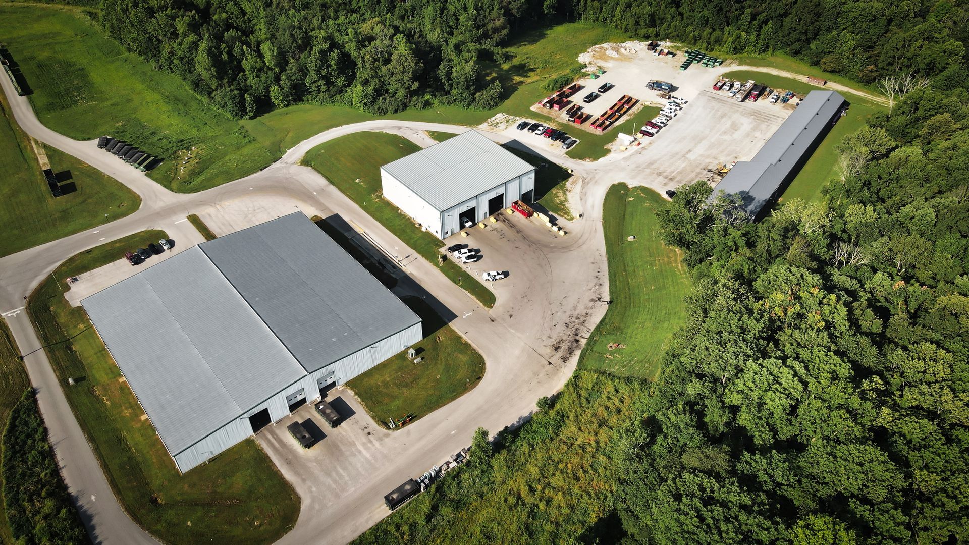 Aerial view of an industrial site with several metal buildings and a parking area surrounded by lush green trees.
