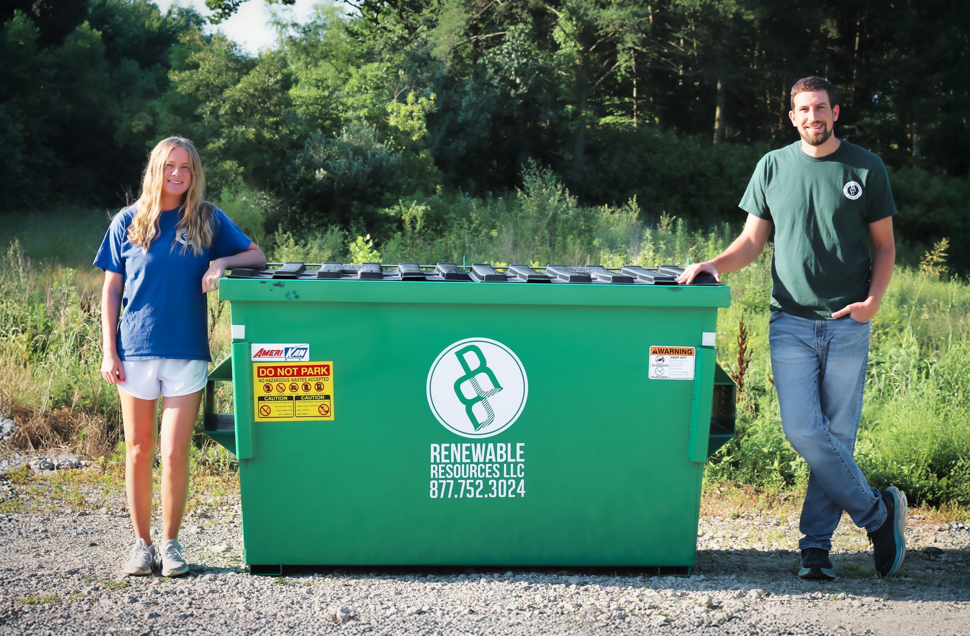 Two people stand beside a large green dumpster in a field. The dumpster has a logo and warning labels.