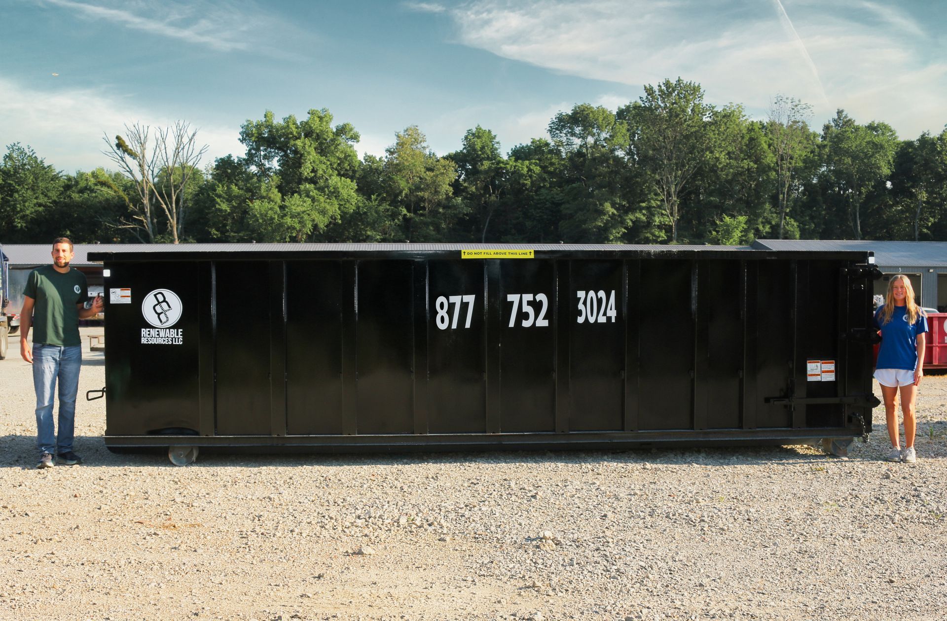 A large black dumpster with an 877-752-3024 phone number, flanked by a man and a girl standing on gravel.