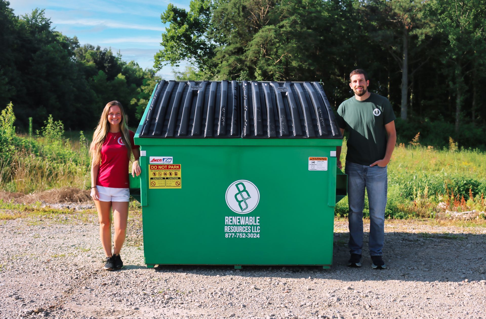 A woman and a man stand beside a large green dumpster in a rural setting.