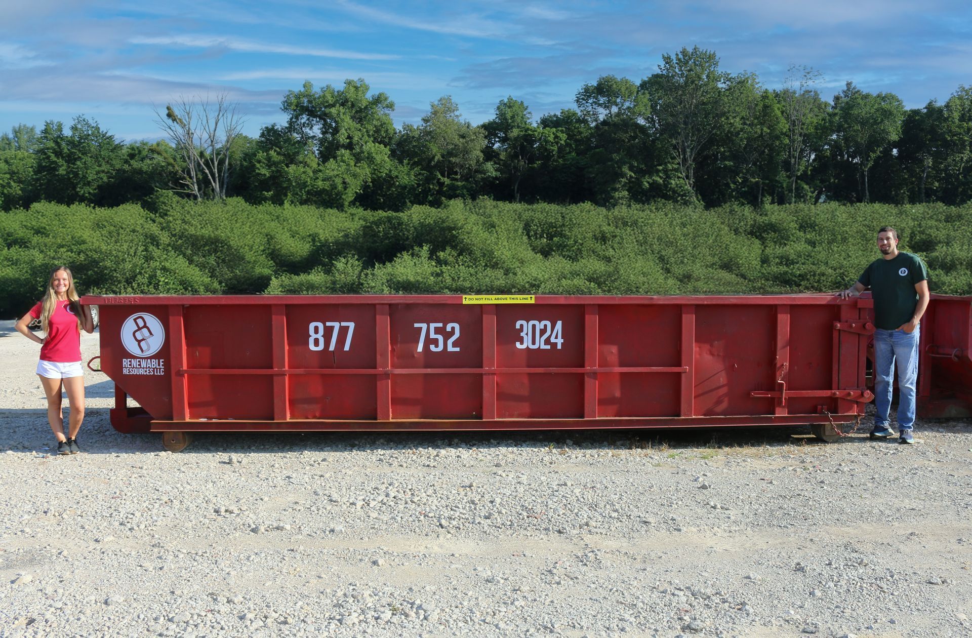 A red dumpster with large numbers on it stands between a woman and a man in a gravel lot.