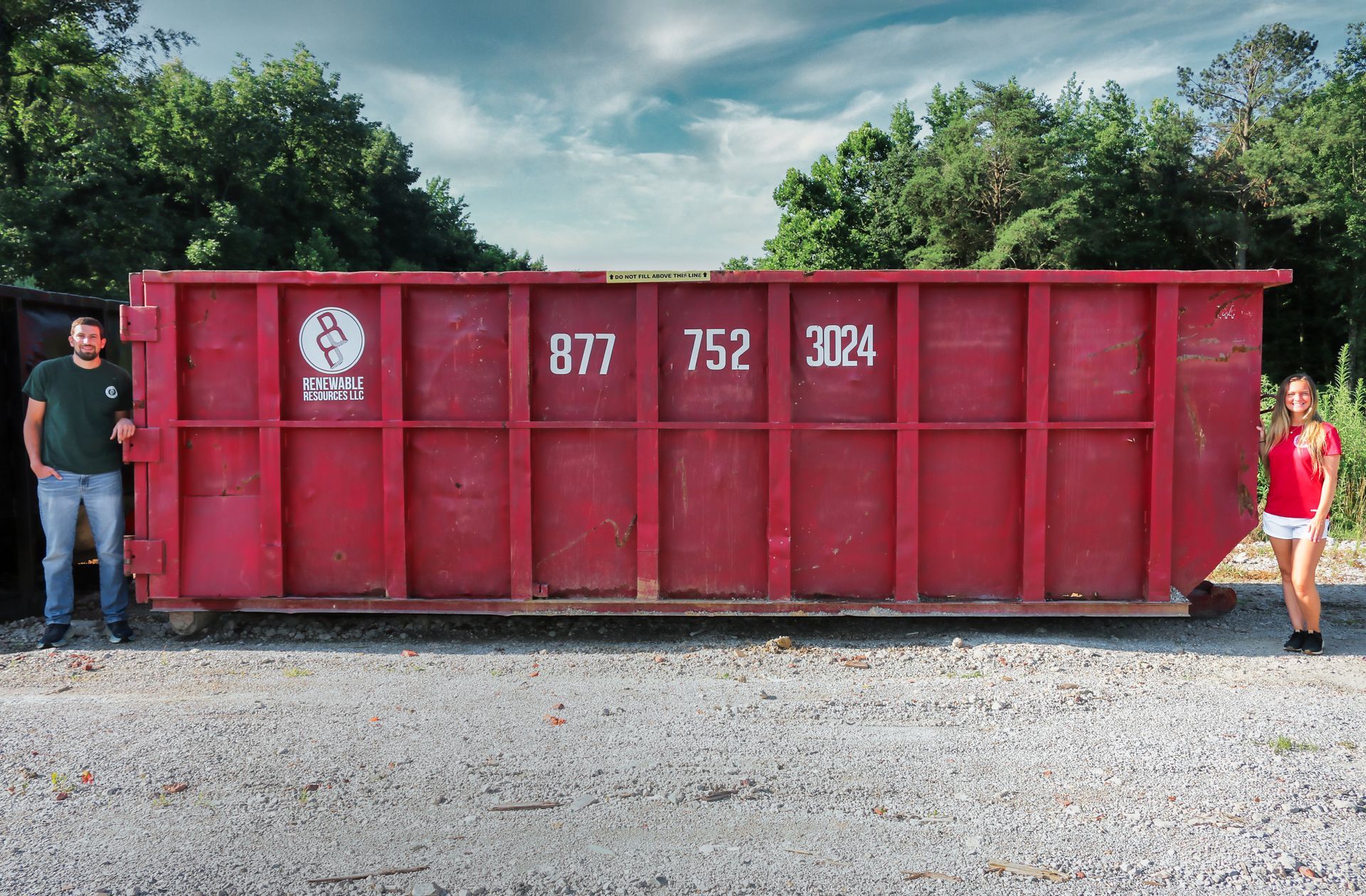 Man and woman stand next to a large, red dumpster. The dumpster has numbers 877 752 3024. Trees in background.