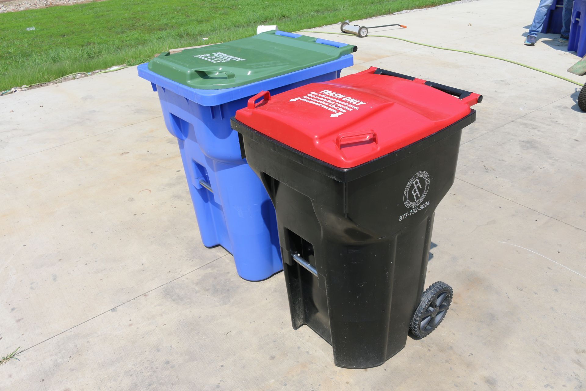 Three wheeled trash bins: blue, black, and a green lid bin. All are on a concrete surface outdoors.