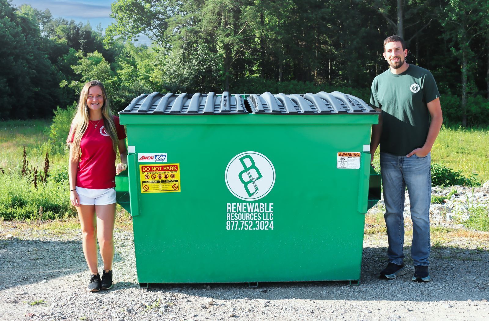 A woman and a man stand on either side of a large green dumpster. They are smiling.