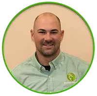 A bald man with a slight smile wears a green collared shirt. He's inside, with a beige background and a green circular frame.