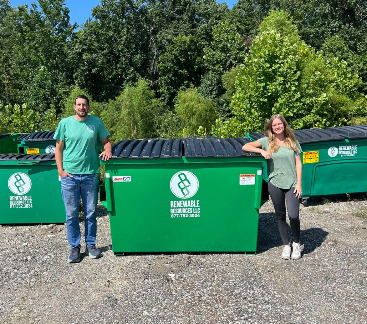 Two people stand beside large green recycling dumpsters in front of a tree line on a sunny day.
