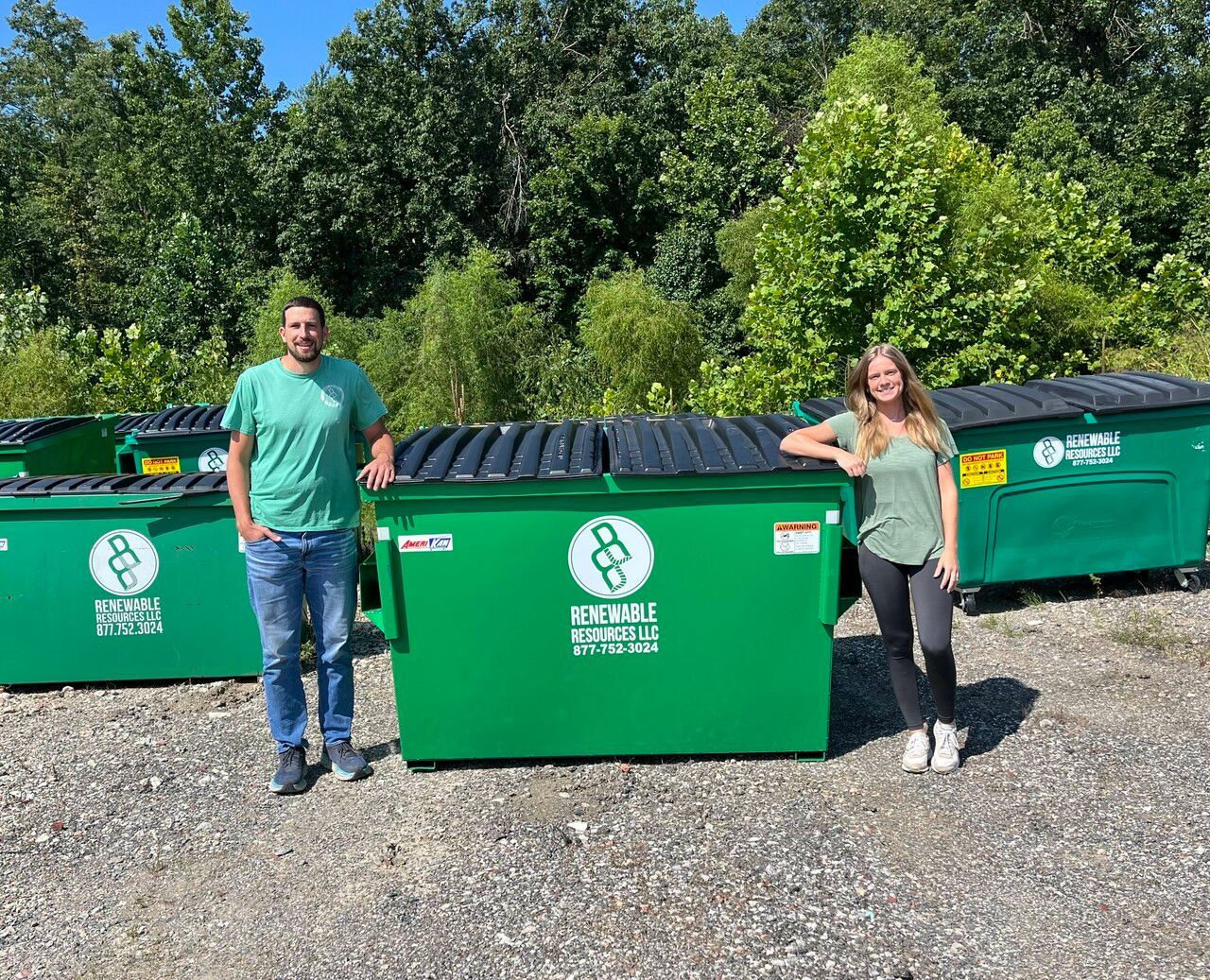 Two people stand beside large green recycling dumpsters in front of a tree line on a sunny day.