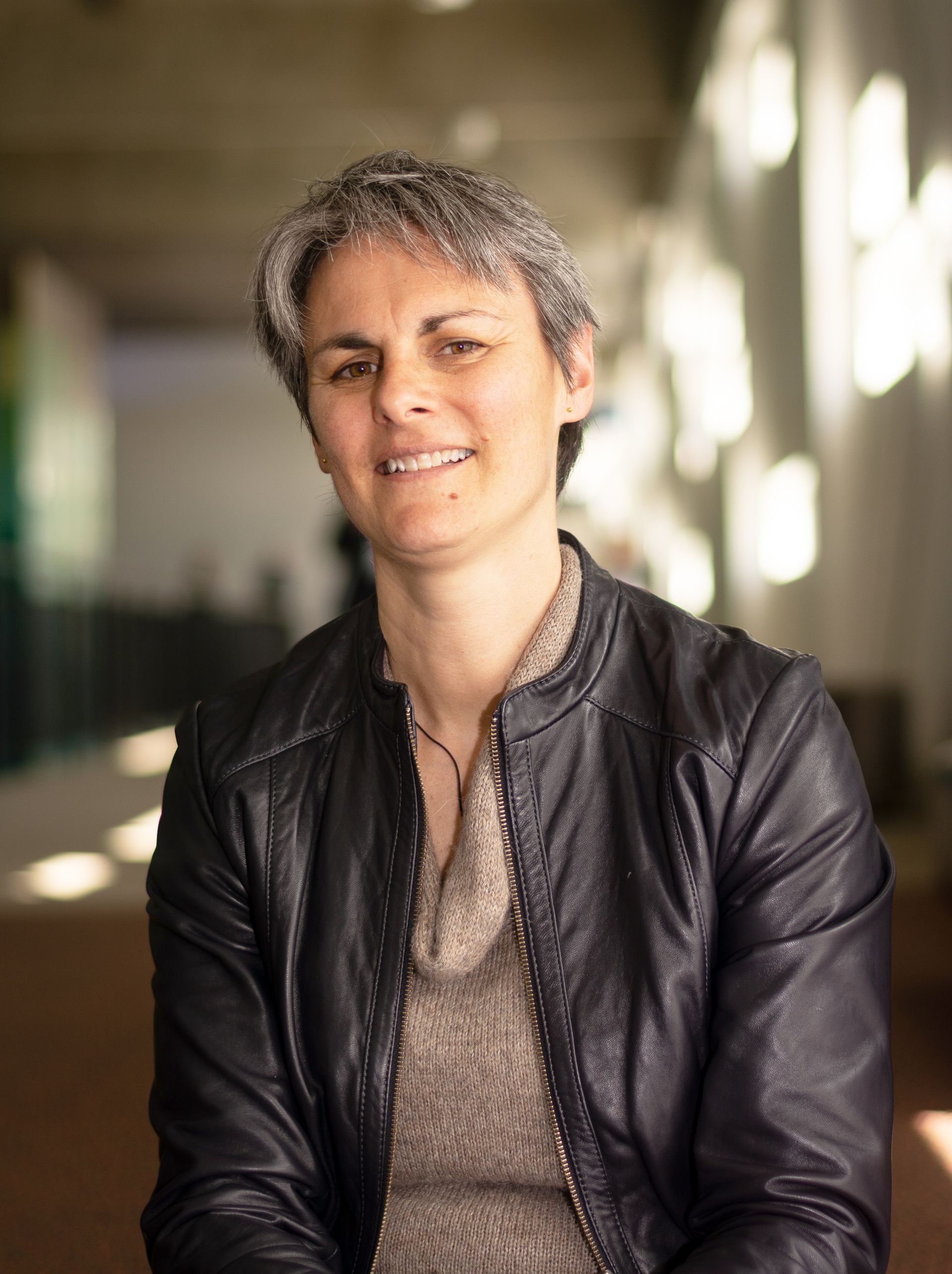 Woman with short gray hair in a black leather jacket smiles indoors.