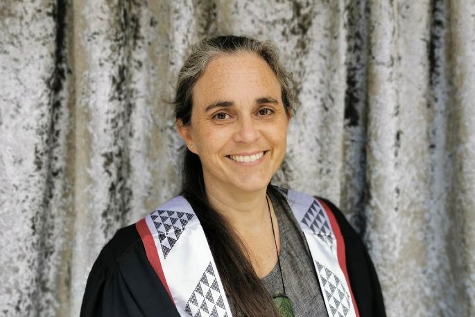 Woman in academic robes smiles, wearing a Māori woven stole, in front of a patterned backdrop.