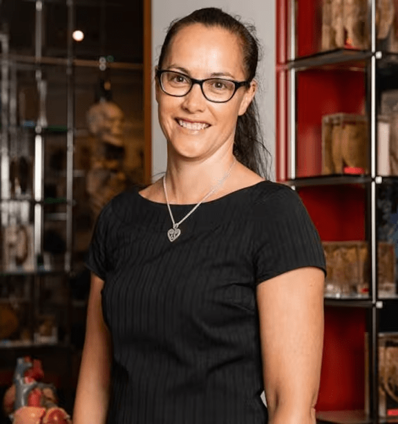Woman with glasses smiles in front of glass-enclosed medical specimens.