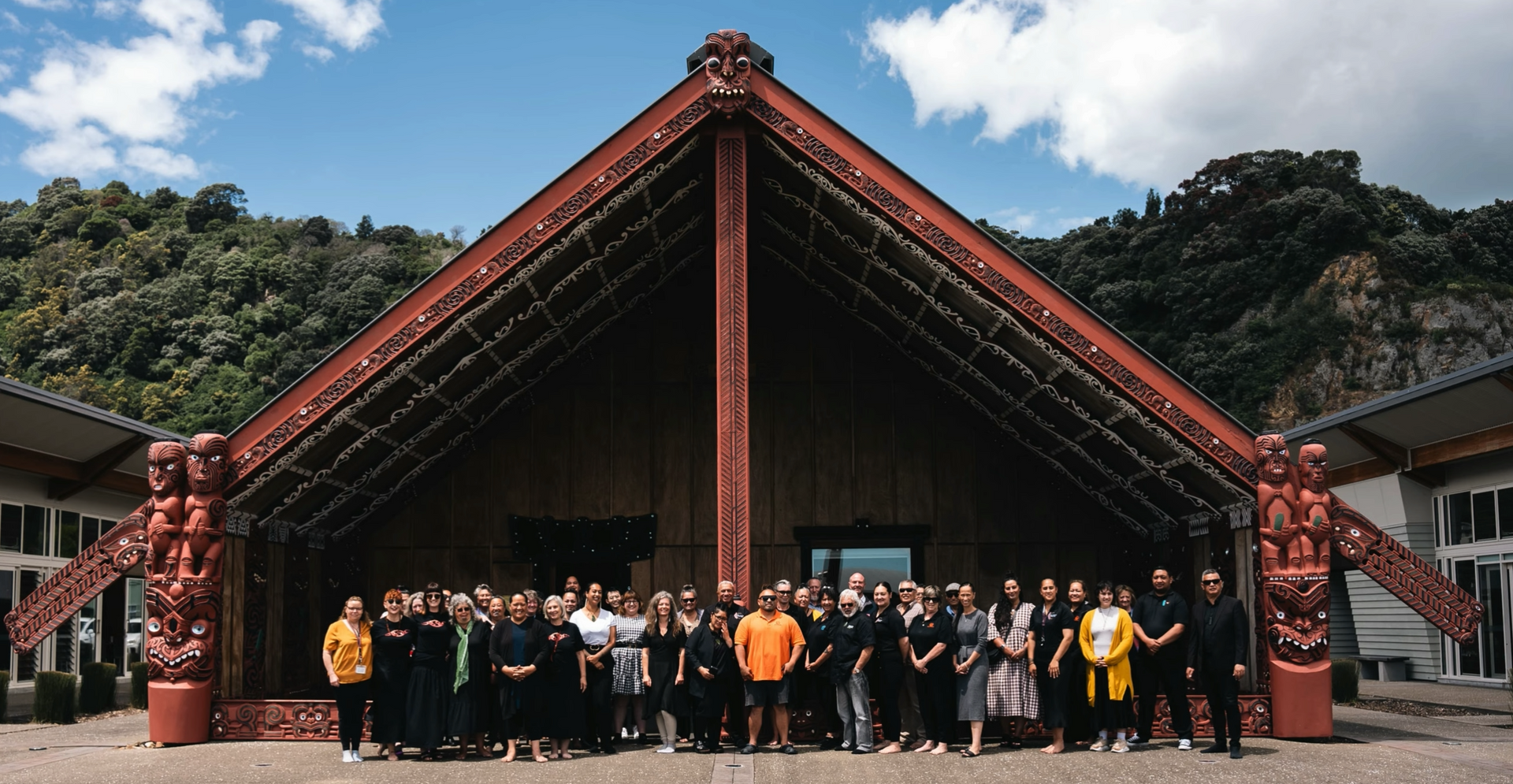 Group of people in front of a Māori meeting house with carved figures and dark red accents, on a sunny day.