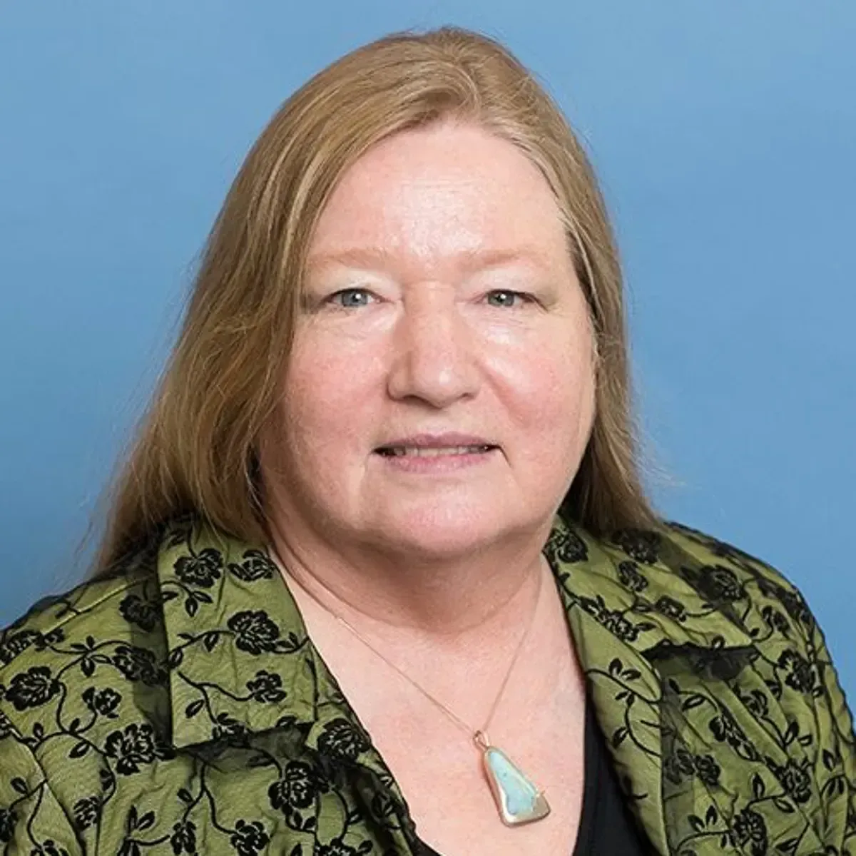 Woman with shoulder-length blonde hair, wearing a green floral-patterned jacket, smiling, blue background.