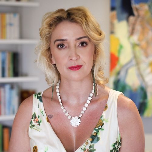 Woman with blonde hair wearing a floral top and necklace, standing in front of a bookshelf and colorful painting.