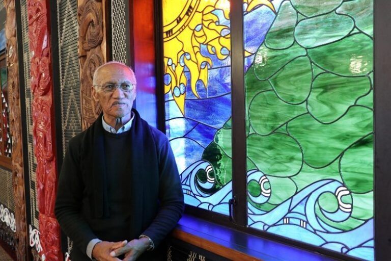 Man stands near a colorful stained-glass window with carved wooden panels visible on left.