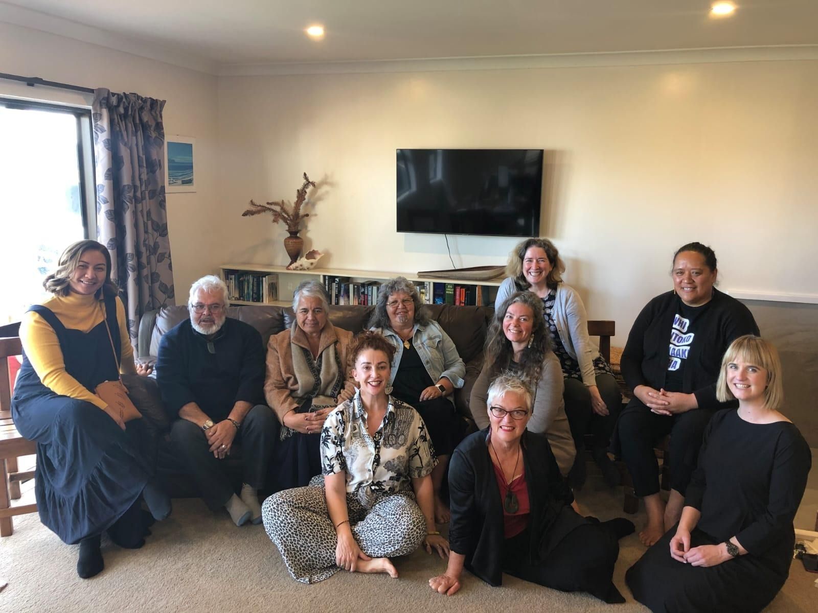 Group of people seated in a living room, smiling at the camera. Neutral background, daytime.