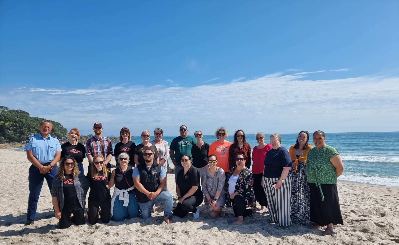 Group of people on a sandy beach posing for a photo under a blue sky, ocean in the background.