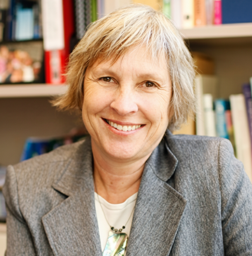 Woman smiling, wearing a gray blazer. Bookshelf visible in background.