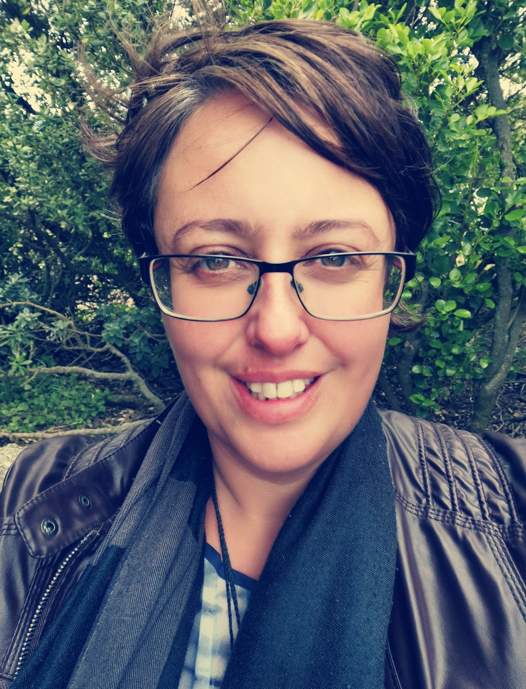 Woman with glasses smiles outdoors, wearing jacket and scarf, trees in the background.