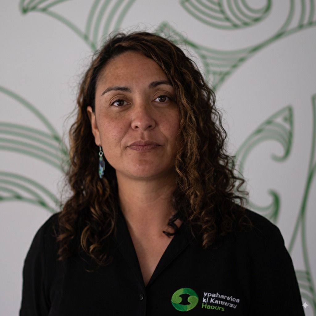 Woman with long wavy brown hair, wearing a black shirt, looking at the camera. A green Māori design is behind her.