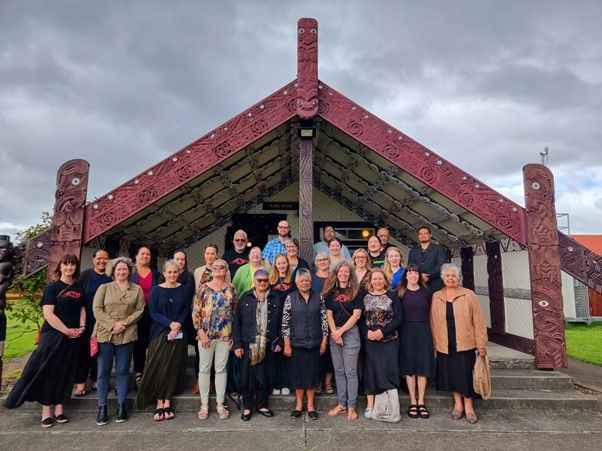 Group of people posing in front of a Māori meeting house with dark red carvings under a cloudy sky.