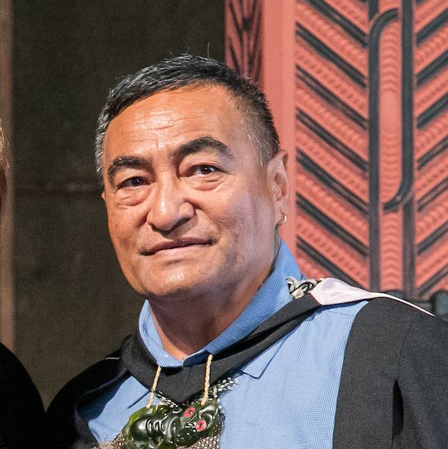 Man wearing a ceremonial cloak and necklace, standing in front of a carved wooden wall.