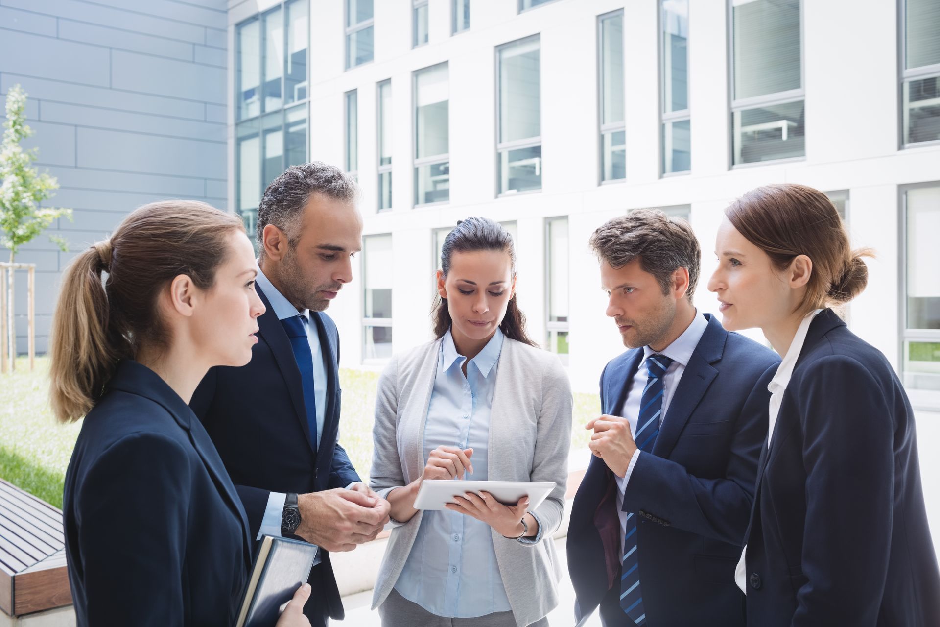 Five businesspeople outside looking at a tablet, discussing. Modern office building in background.