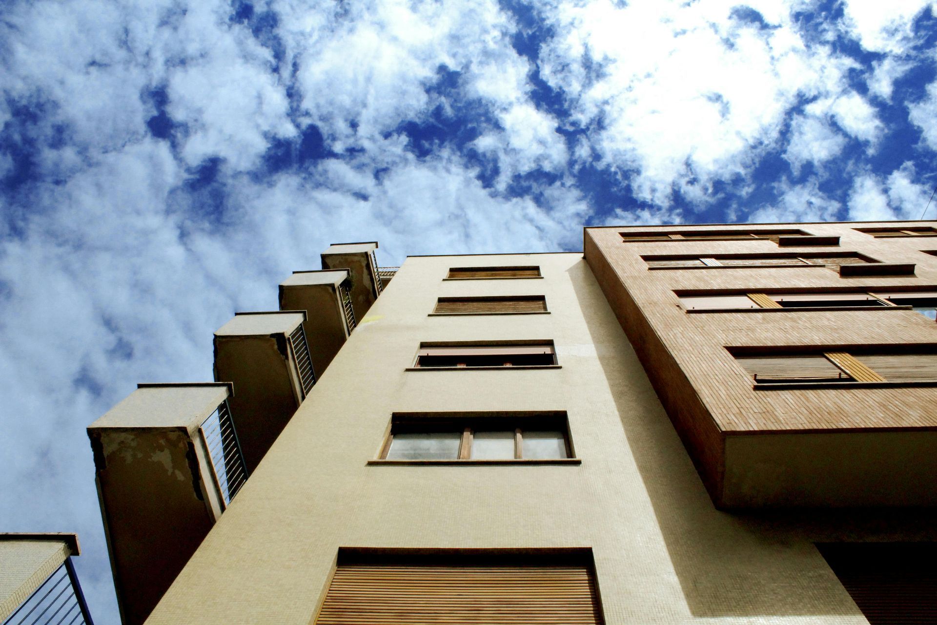 Low-angle view of a beige and brown building against a bright blue sky with scattered clouds.
