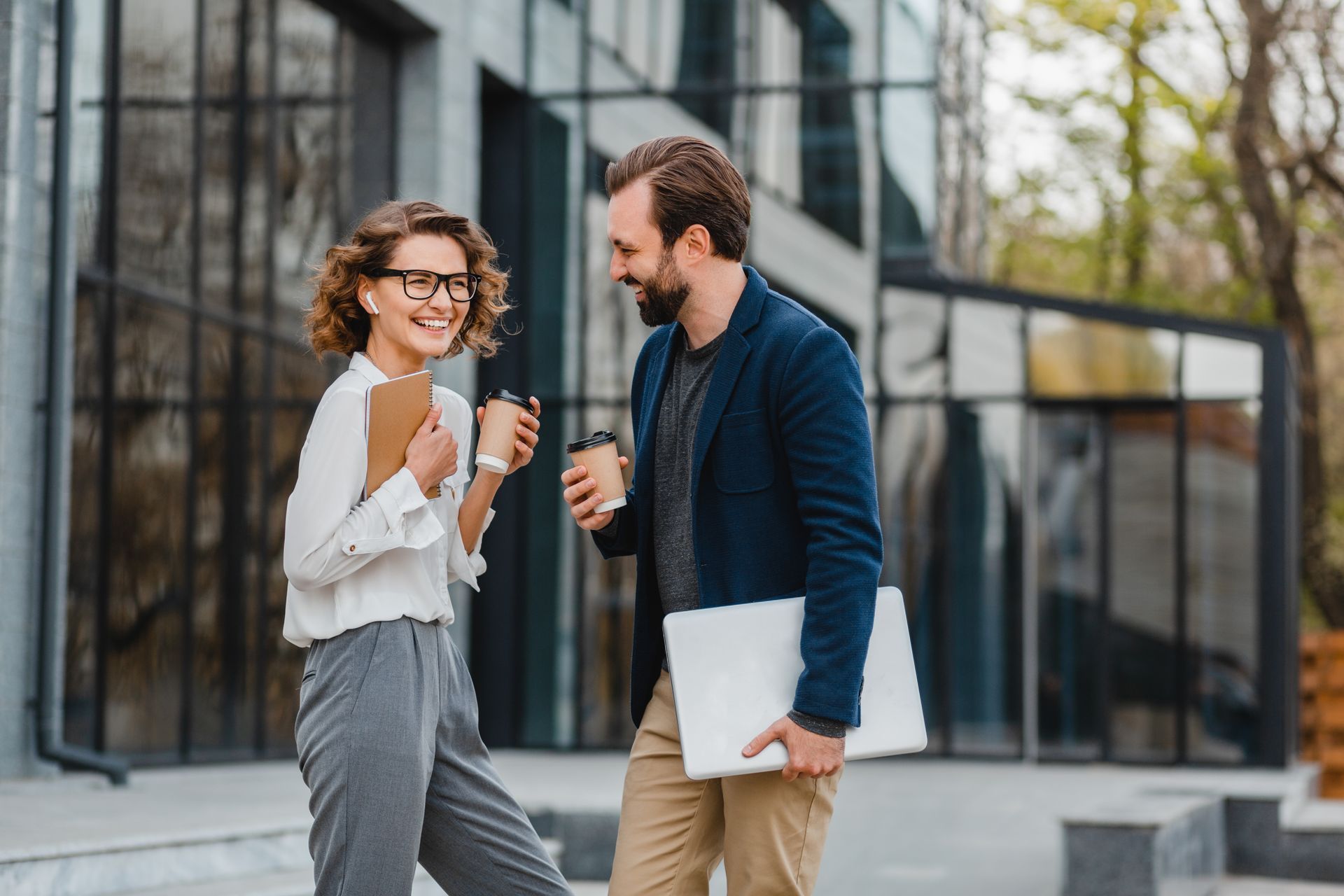 Woman with glasses and man with beard laugh while talking outdoors, holding coffee and a laptop