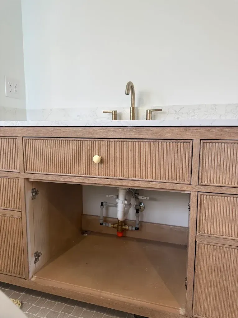 Wooden bathroom vanity with gold faucet, unfinished cabinets, and exposed plumbing.