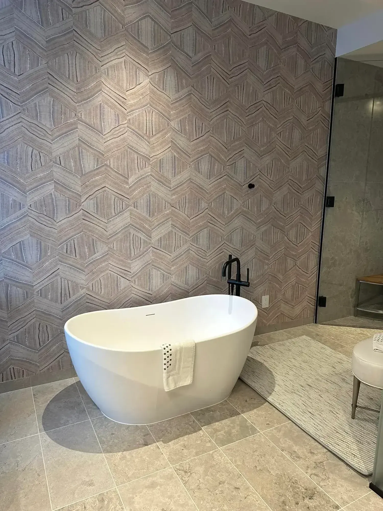 Modern bathroom with a white oval bathtub, patterned tile accent wall, and stone floor.