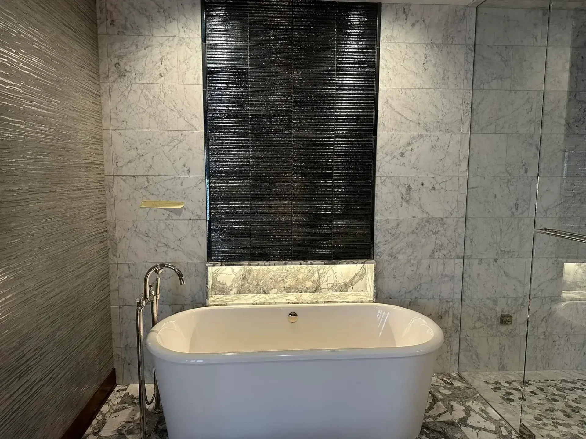 Freestanding white bathtub in a modern bathroom with marble walls and a black tiled accent.