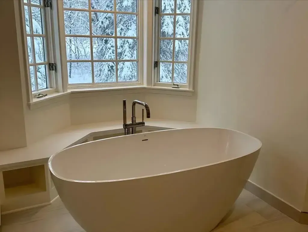 White freestanding bathtub beneath a large window. Neutral-toned bathroom.