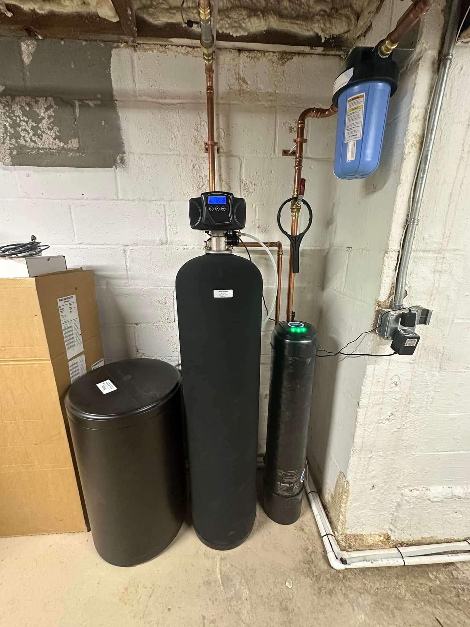 Water filtration system in a basement. Includes a water softener tank, filter, and brine tank, with copper pipes against a concrete wall.
