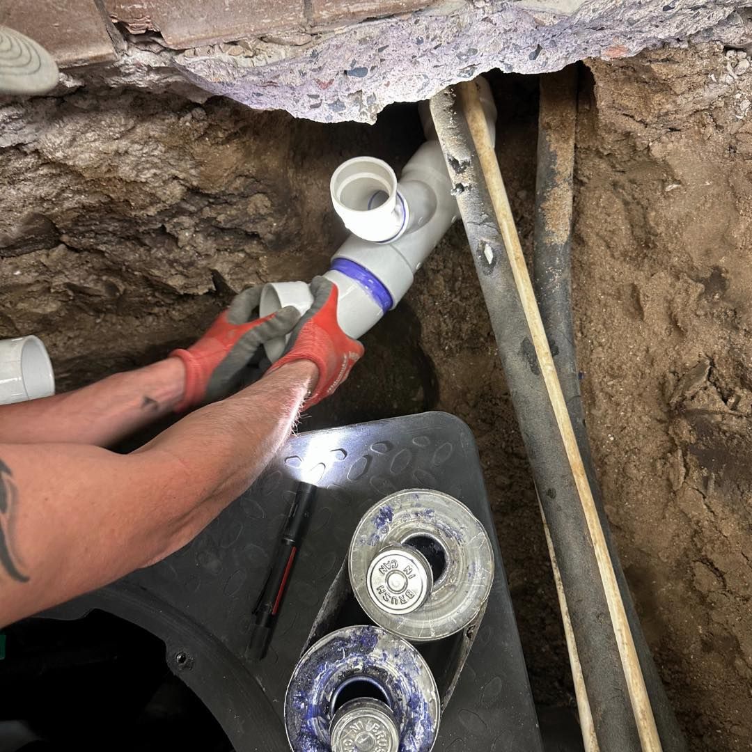 A person with red gloves is connecting white PVC pipes underground next to a brick wall.