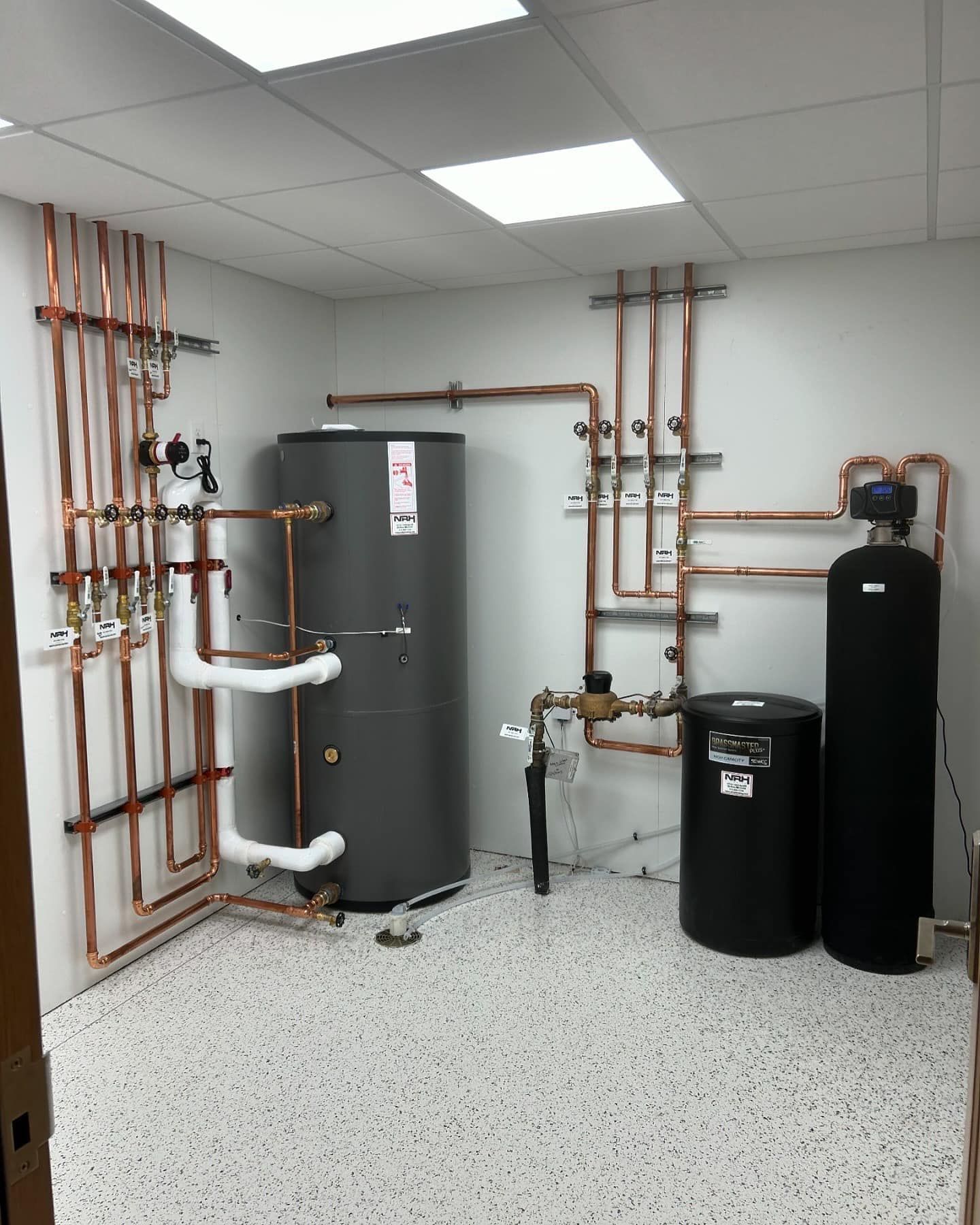 Copper pipes, water heater, water softener, and tanks in a utility room with white walls and speckled floor.