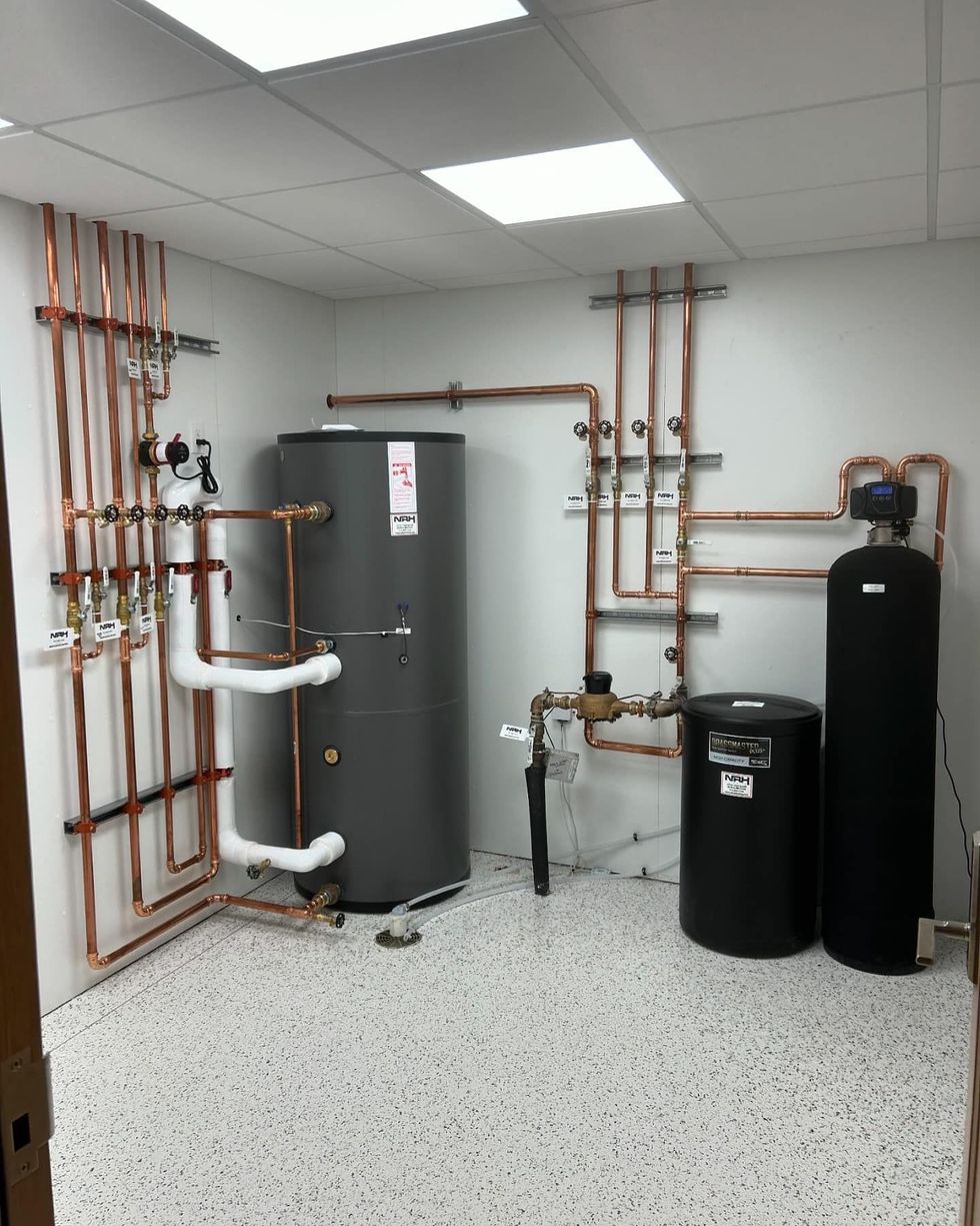 Copper pipes, water heater, water softener, and tanks in a utility room with white walls and speckled floor.