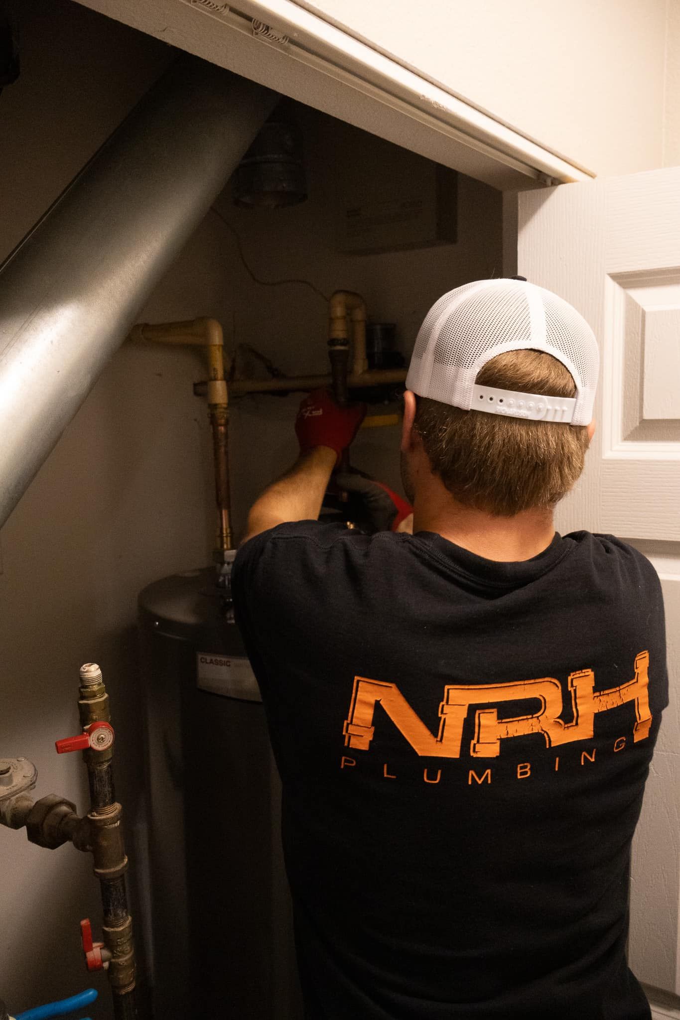 Person in black shirt and white hat working on a water heater in a closet.