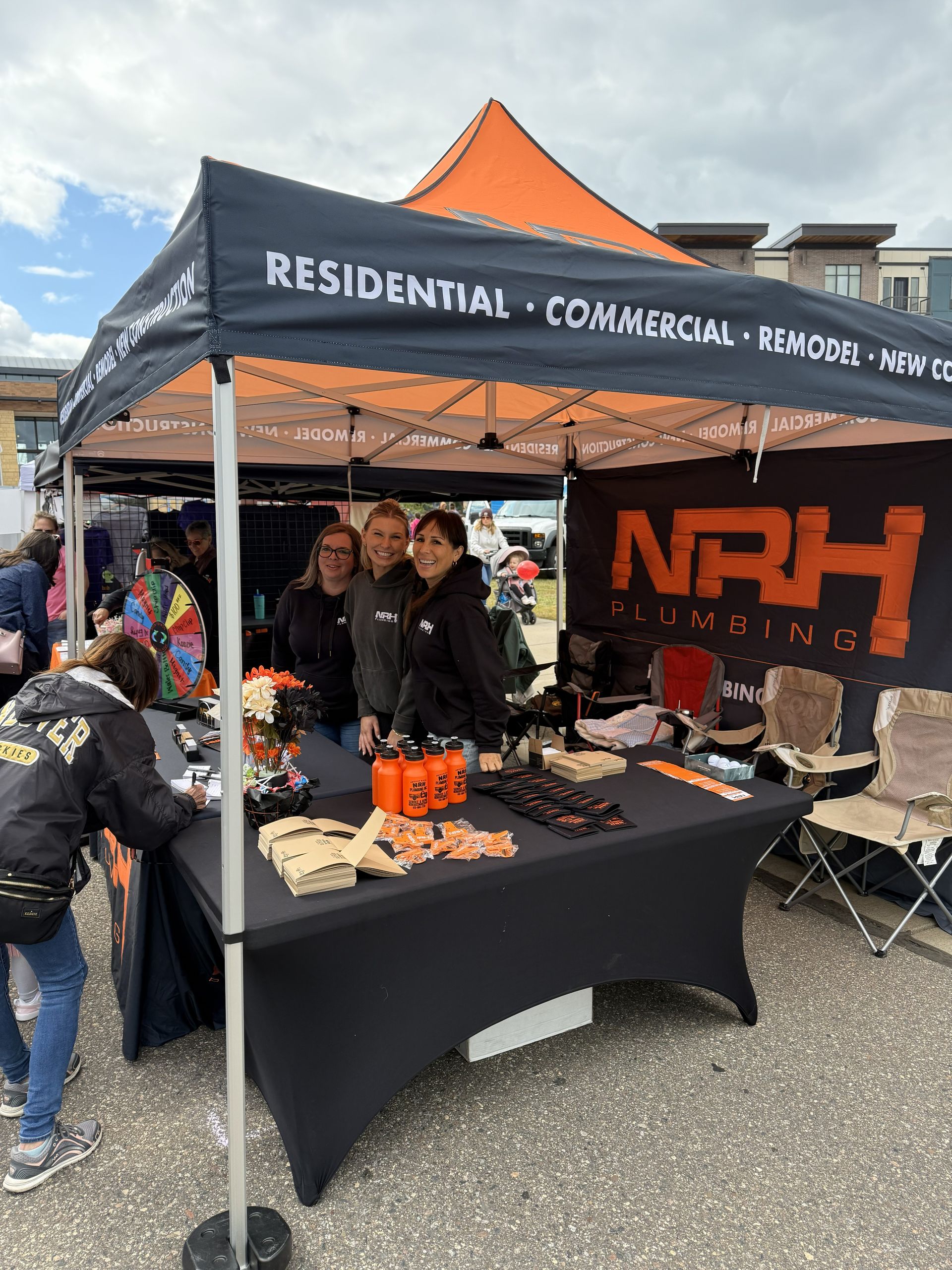 NRH Plumbing booth at an outdoor event, staffed by three women, offering treats and information.