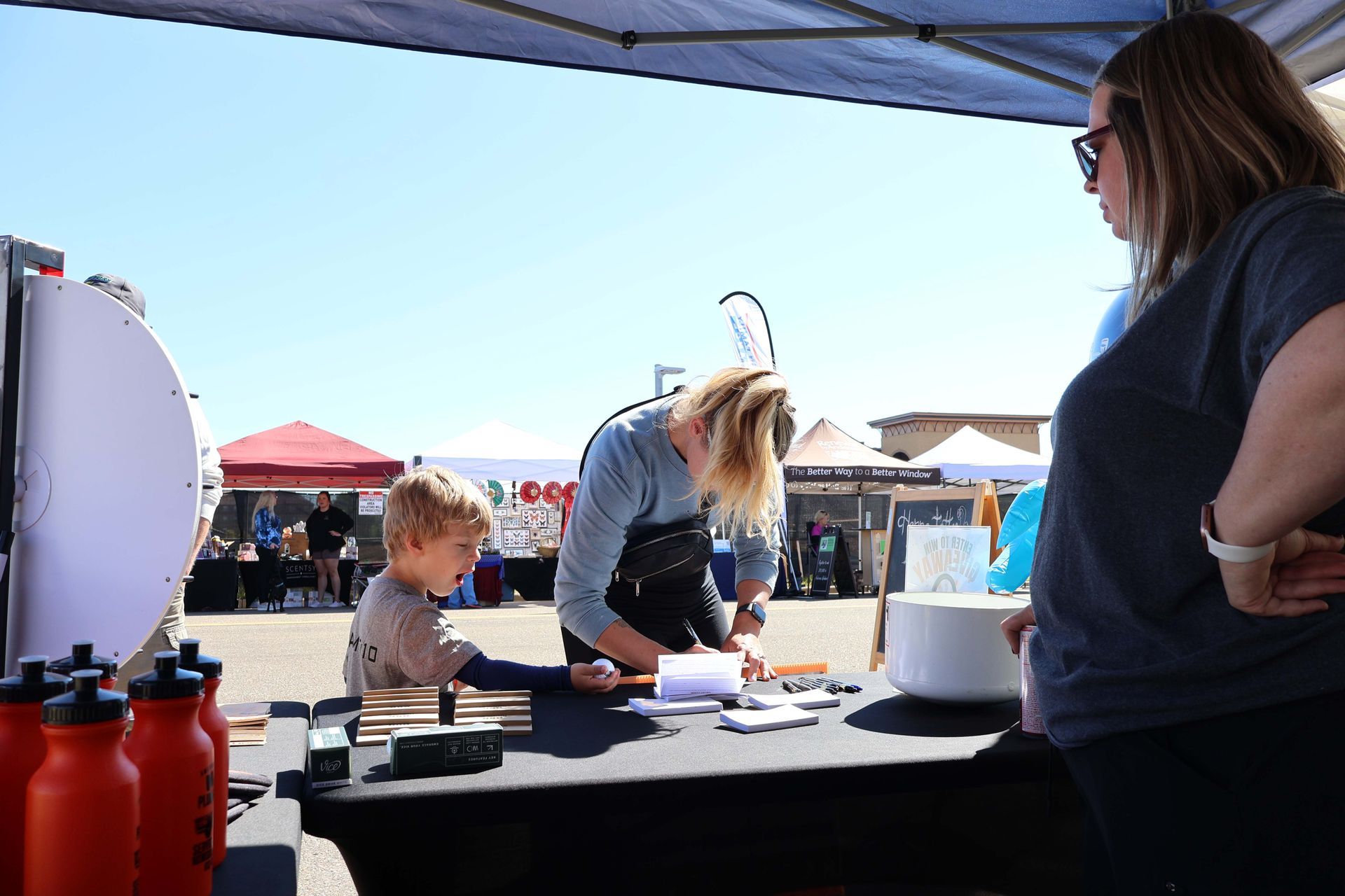 Child getting an imprint at a booth under a canopy, with an adult helping. Outdoors on a sunny day.