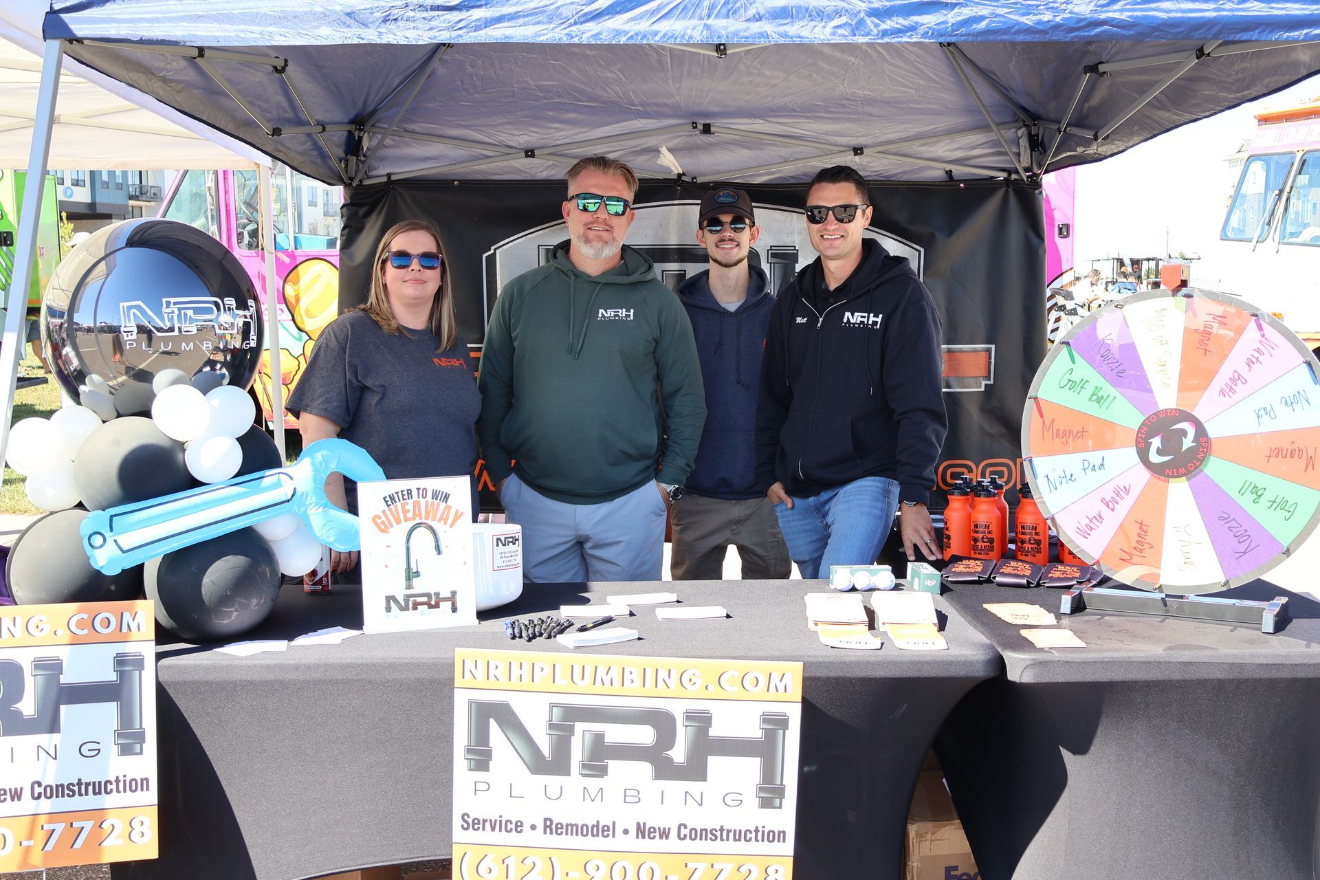 Four people at an NRH Plumbing booth under a tent, smiling. There's a prize wheel and balloon art.