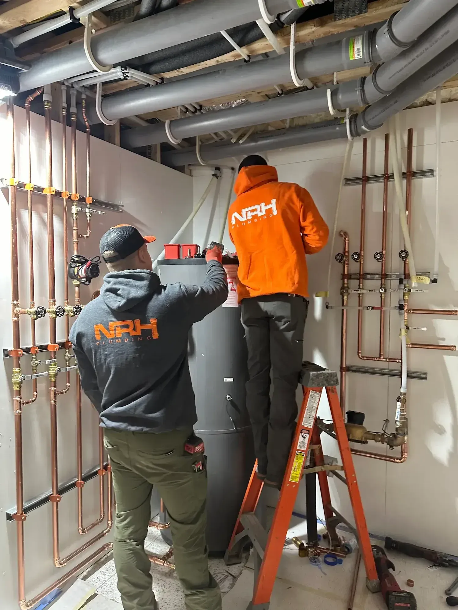Two workers in orange and gray shirts install plumbing near a black water heater in a basement.