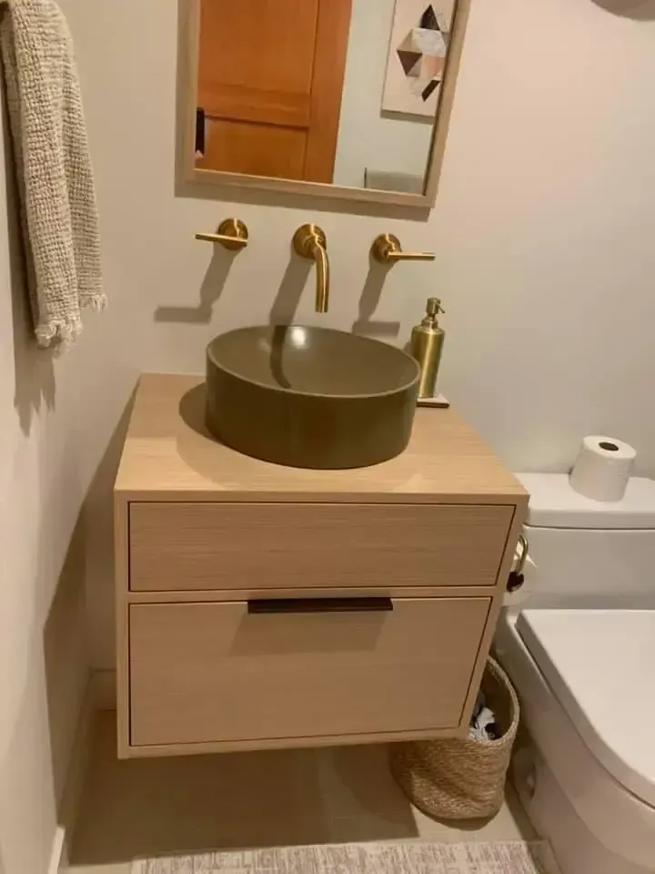 Modern bathroom with a vessel sink on a light wood cabinet. Gold faucet and mirror frame. Beige walls, toilet on the right.