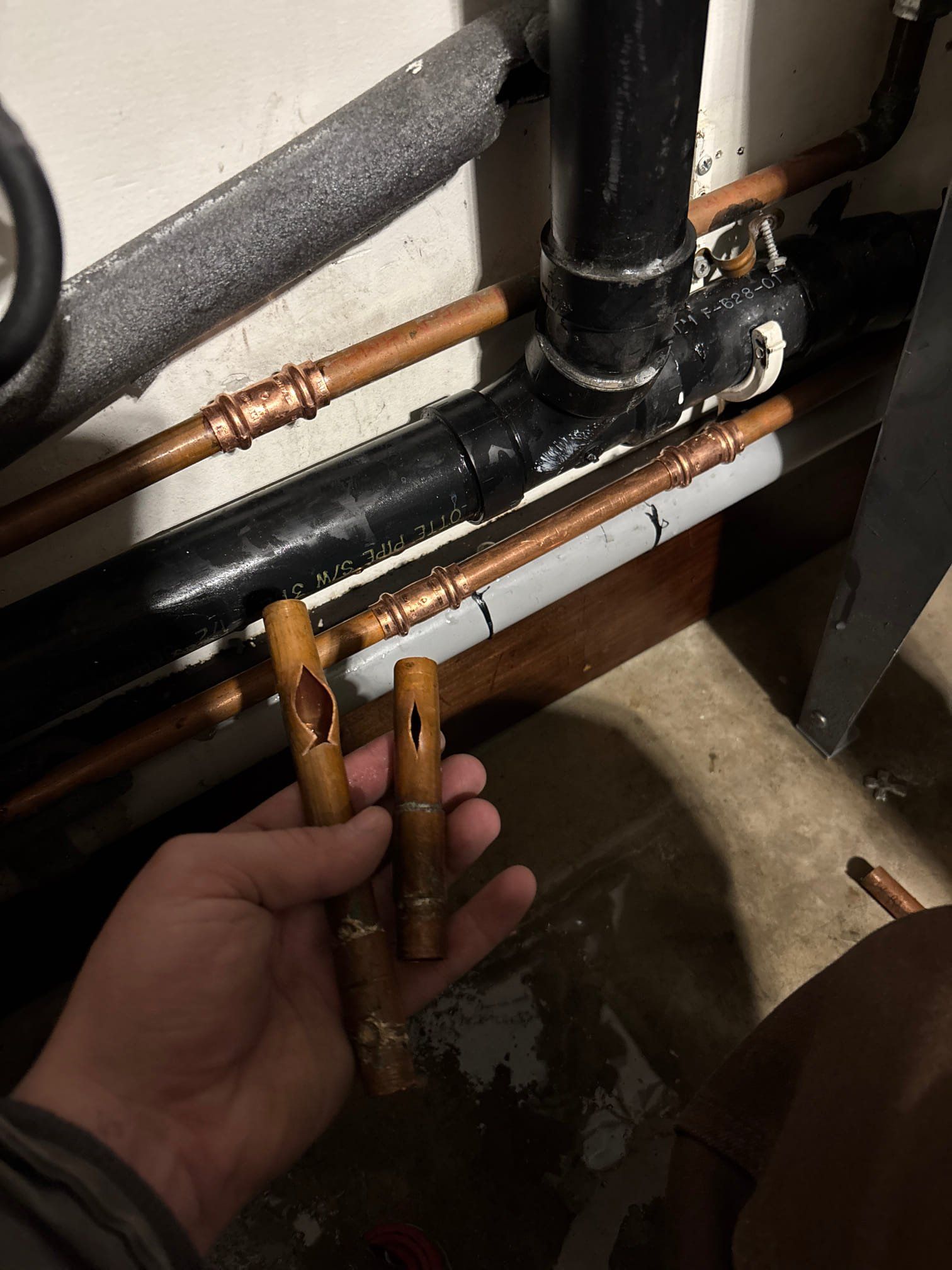 Two people working on plumbing in a room. One applies sealant, tools and a propane tank are present.