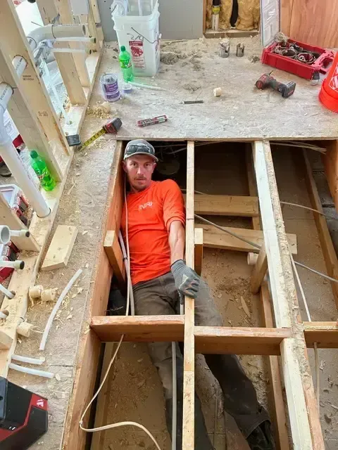 Man in orange shirt inside a floor frame, plumbing and construction materials around.