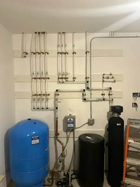 Water pipes and tanks mounted on a white wall. Includes a blue tank, black tanks, and copper/plastic pipes.