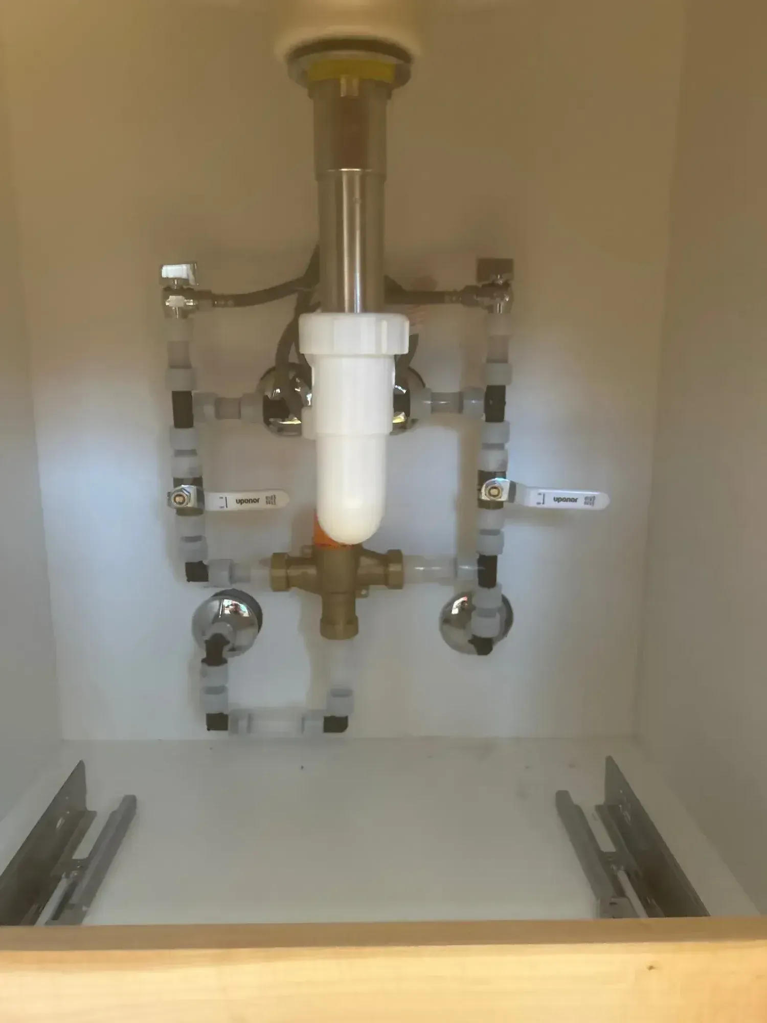 Plumbing pipes and fixtures under a bathroom sink. White and clear plastic piping, chrome fixtures.