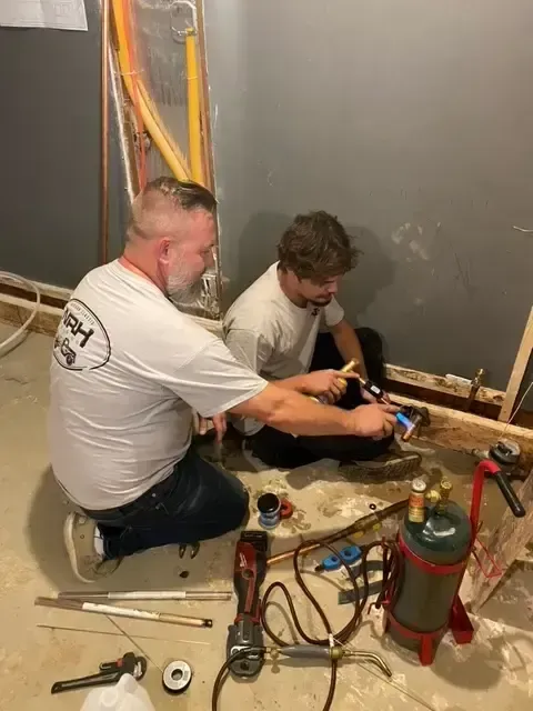 Two people working on plumbing in a room. One applies sealant, tools and a propane tank are present.