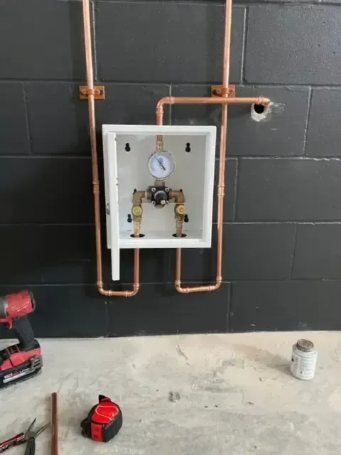 Copper pipes and valves installed inside a white box on a black tiled wall, tools in foreground.